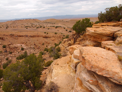 [Near the top of a rock outcropping with stacked horizontal slabs of rock. This view is at least several hundred feet above the lower levels. While there is some green shrubs growing on the rock, the ground below is nearly all brown dirt and rock.]